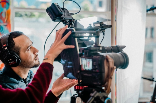 A person operating a video camera, while another person observes A person wearing a red long-sleeved shirt operates a video camera, while another person observes the scene being filmed